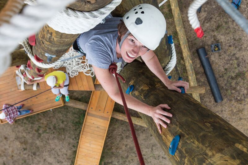 The image shows a person wearing a helmet and climbing a wooden structure. They are using a rope for support and appear to be enjoying the activity. Other people are visible in the background, also engaged in climbing. The perspective is from above, offering a clear view of the climber and the surrounding environment.